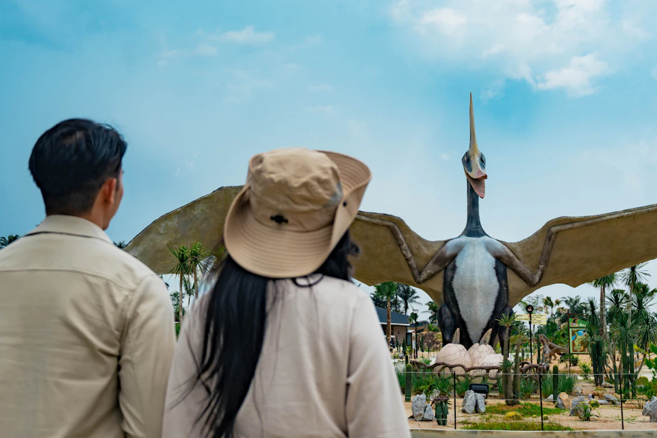 Two people facing a large pterosaur statue in a park under a blue sky.