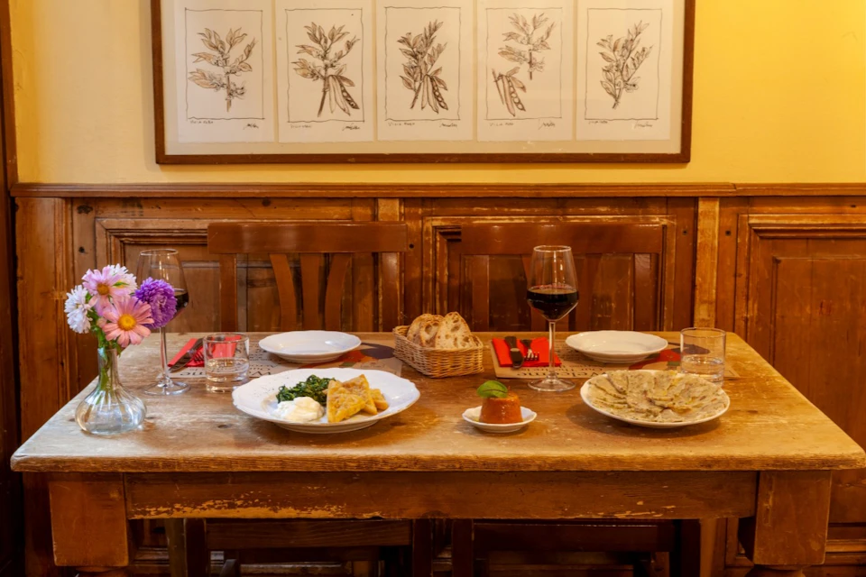 A rustic dining table set with plates of food, red napkins, a glass of wine, and a flower vase, against a wall with botanical drawings.