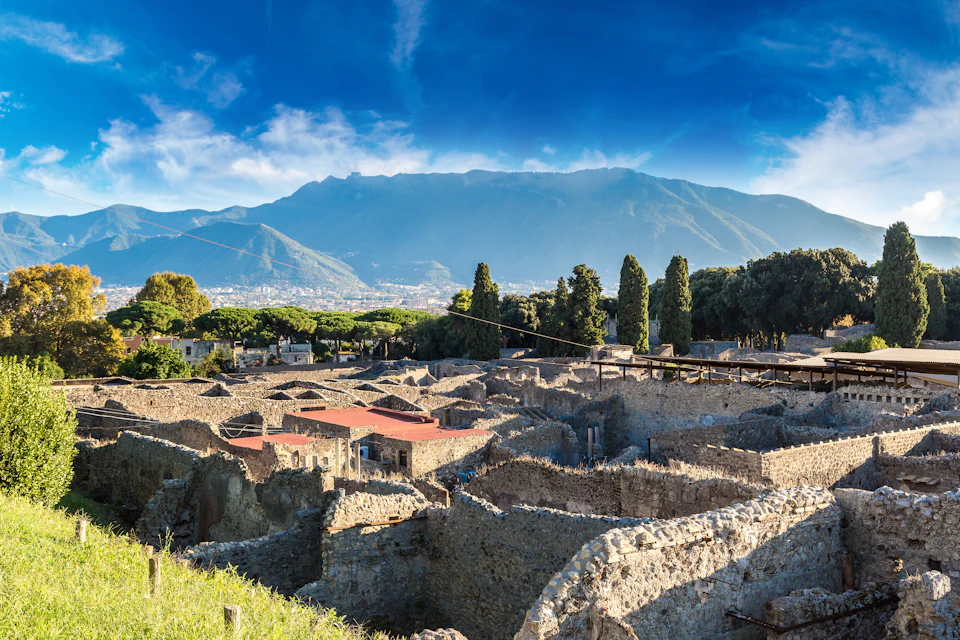 Ancient ruins with stone walls and red roofs, surrounded by trees, with mountains and a blue sky in the background.