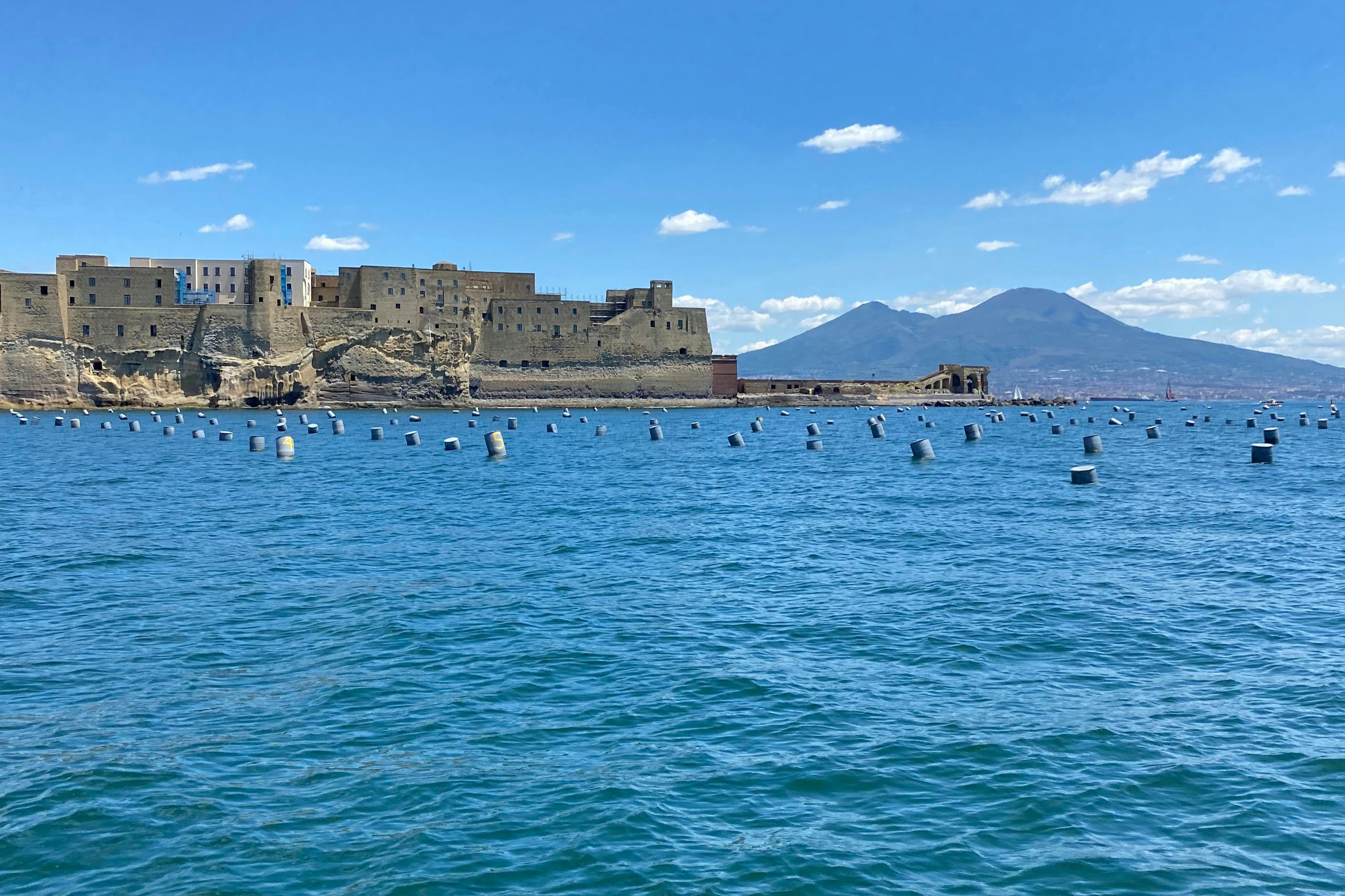 Castillo sobre una formación rocosa costera con numerosas boyas en primer plano y montañas al fondo bajo un cielo azul despejado.