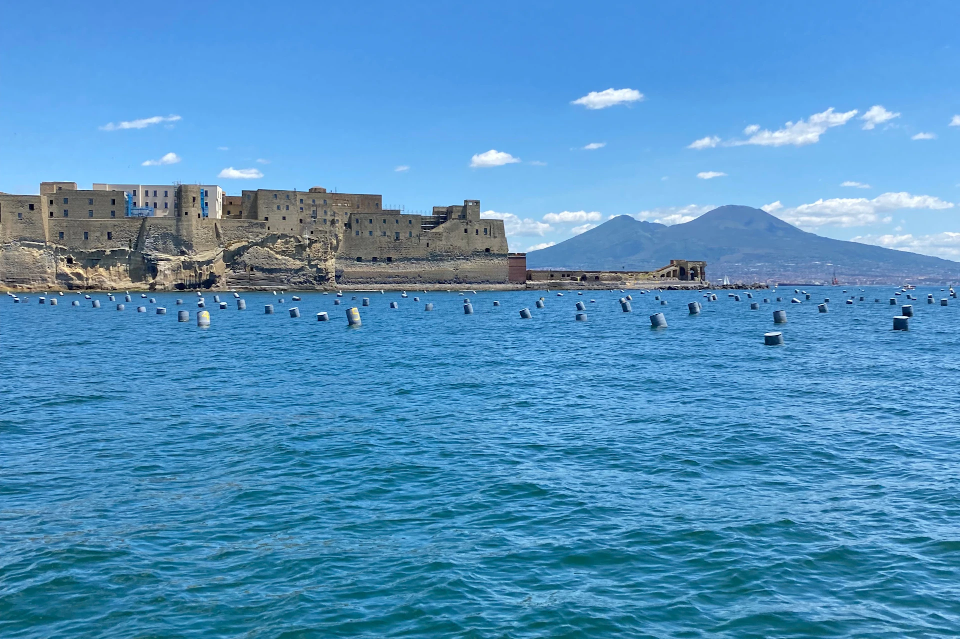 Castle on a coastal rock formation with numerous buoys in the foreground, and mountains in the background under a clear blue sky.