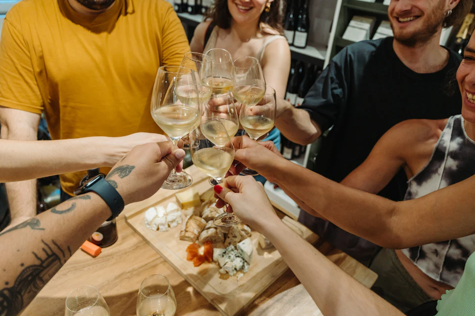 A group of people toasting with wine glasses over a table with assorted cheese, bread, and other foods.