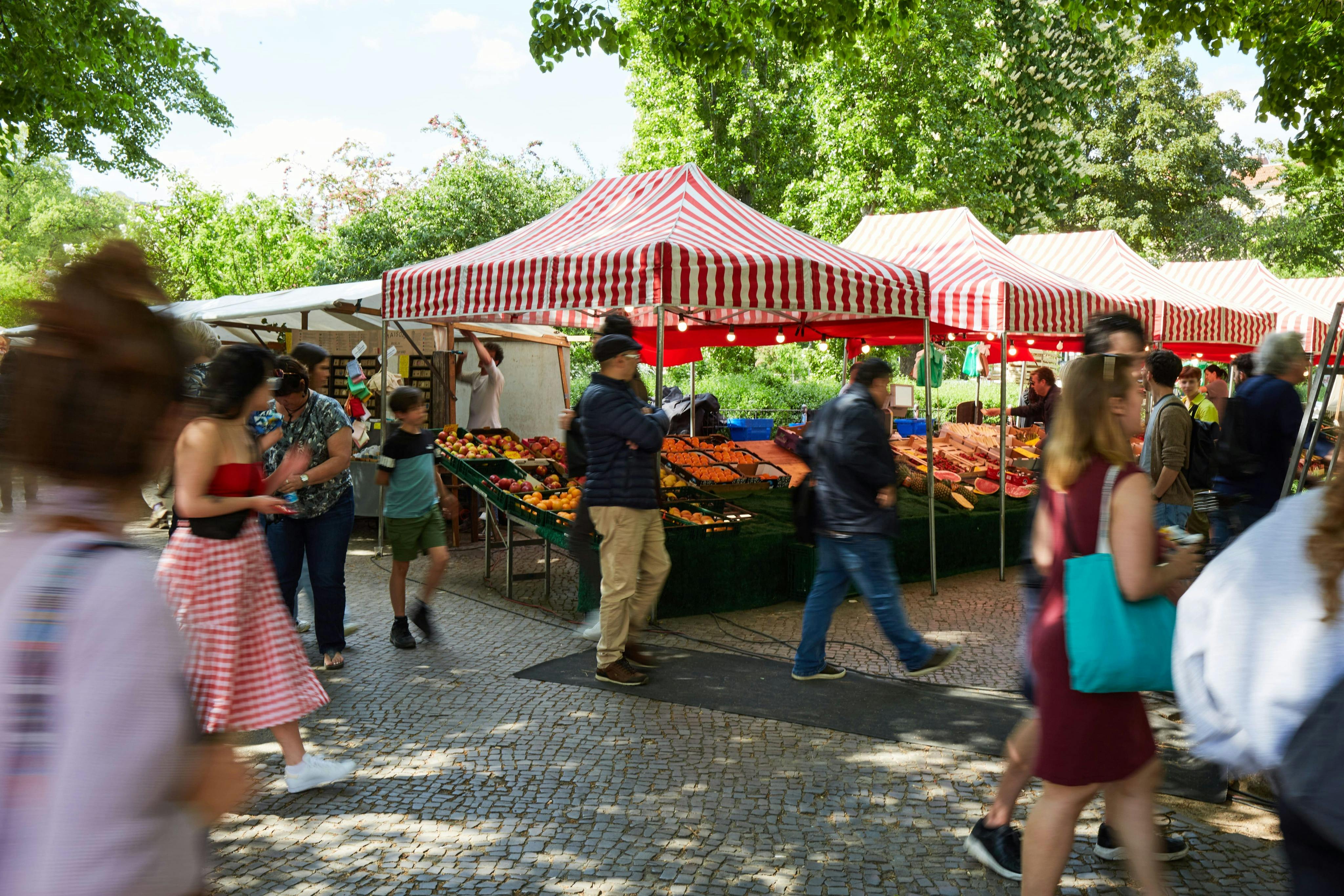 Πάγκοι στην υπαίθρια αγορά με κόκκινα και λευκά ριγέ στέγαστρα που πωλούν φρούτα και λαχανικά, περιτριγυρισμένοι από ανθρώπους που περιηγούνται.