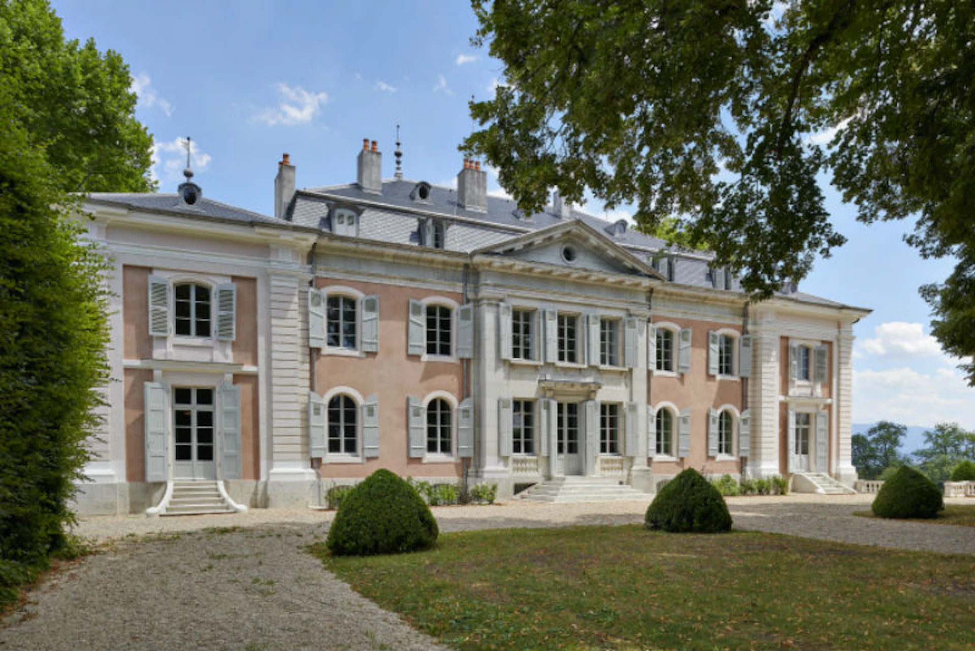 A large, elegant mansion with pink and white facade, multiple chimneys, and neatly trimmed bushes, under a blue sky.