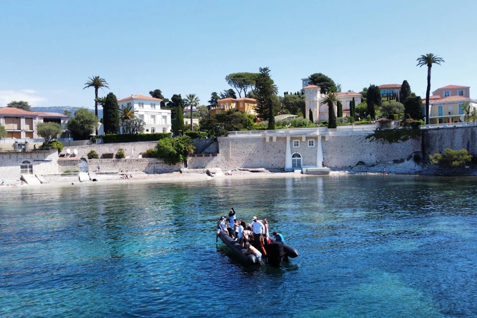 A group of people on a small inflatable boat in clear blue water near a scenic coastal area with villas and trees.