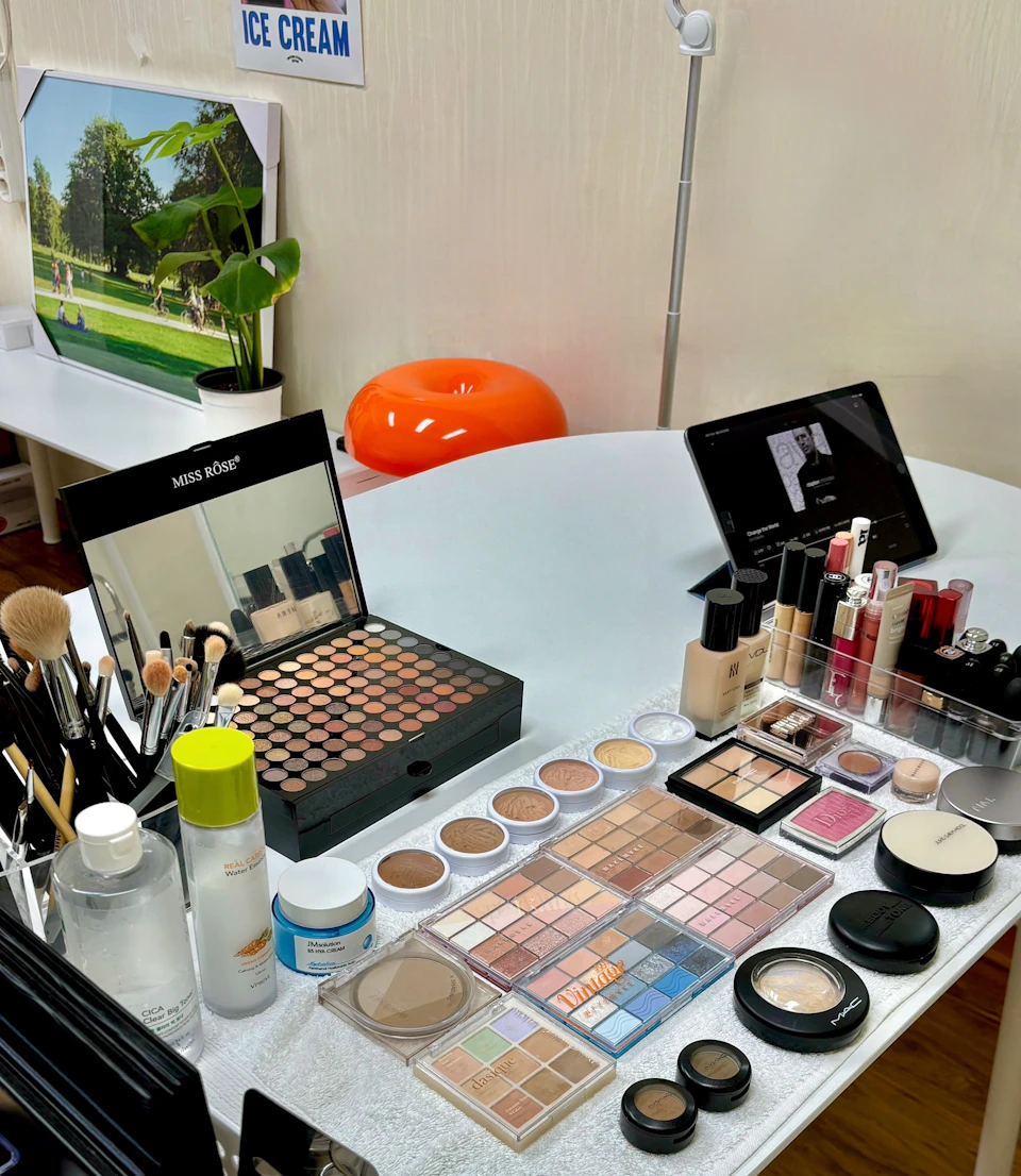 A table with various makeup products, brushes, and palettes arranged neatly. A mirror and digital devices are also present.