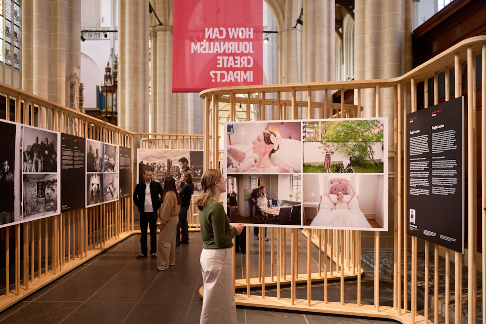 An indoor exhibition featuring photographs displayed in wooden frames. Visitors are looking at the displays. A pink banner hangs above.