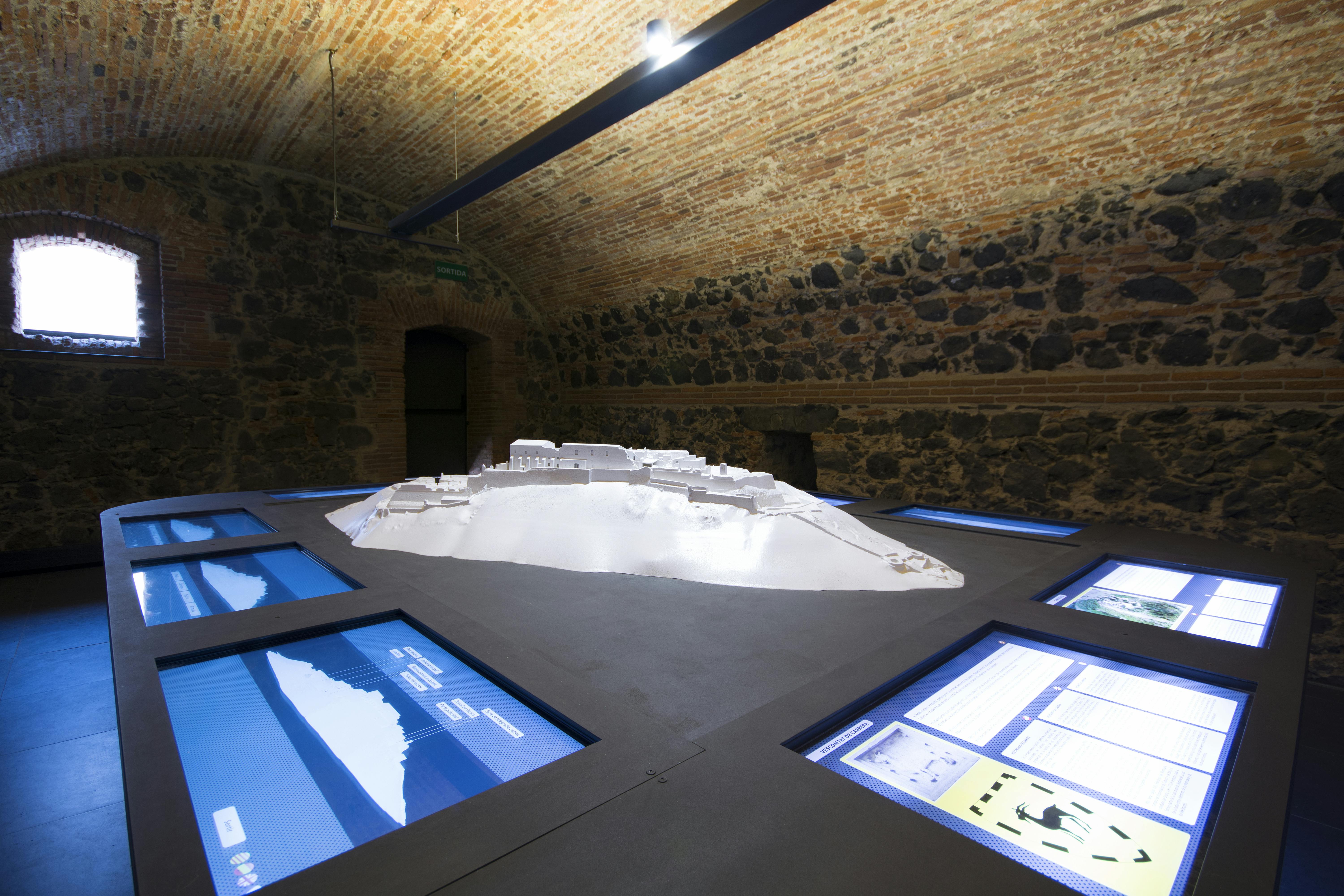 A room with brick and stone walls featuring a central 3D model display surrounded by multiple interactive screens.