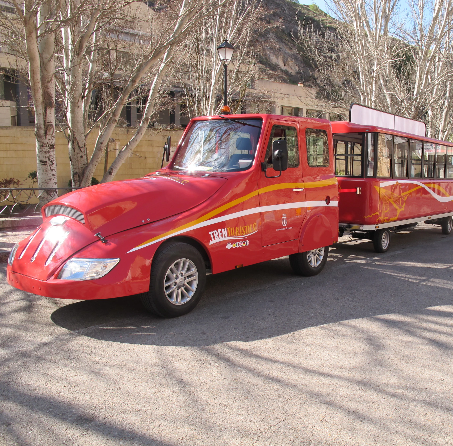 A bright red tourist train with a cabin and open passenger carriages on a street lined with leafless trees.