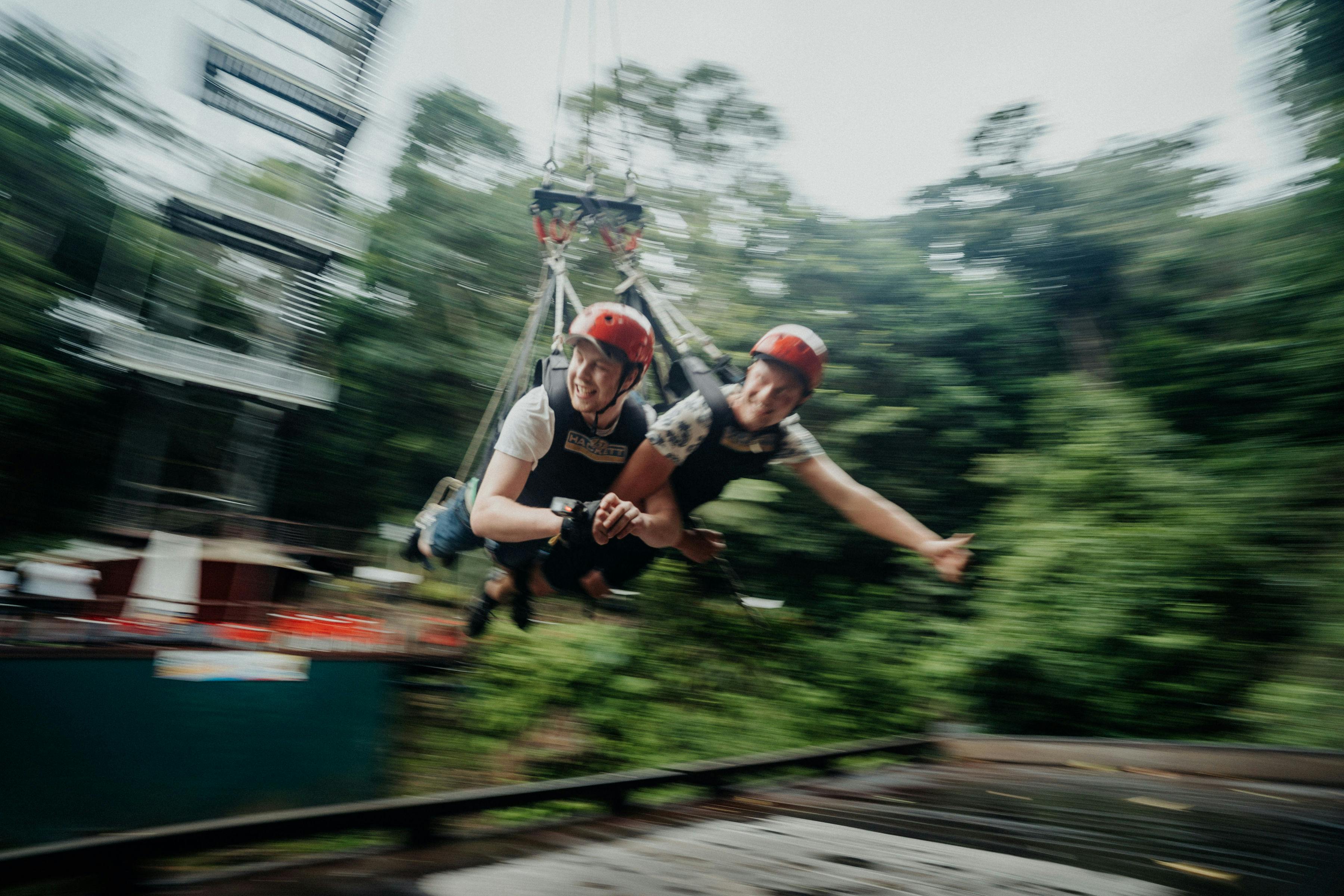 两个人戴着头盔和马具，在户外探险公园的滑索上悬挂在半空中。 树木在背景中模糊不清。