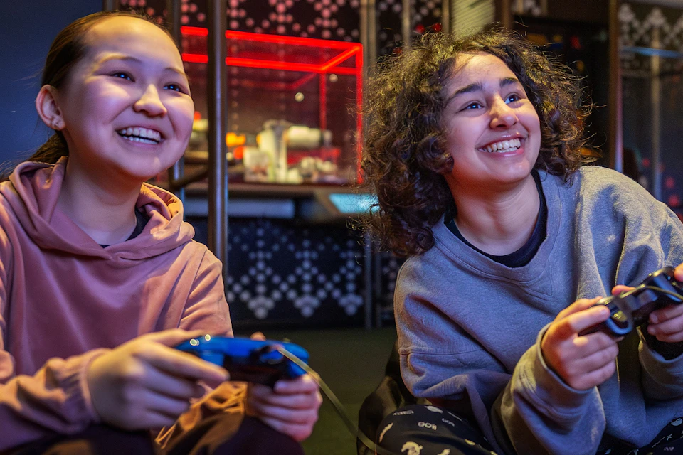 Two individuals sitting indoors, smiling and playing a video game. One is holding a blue controller.