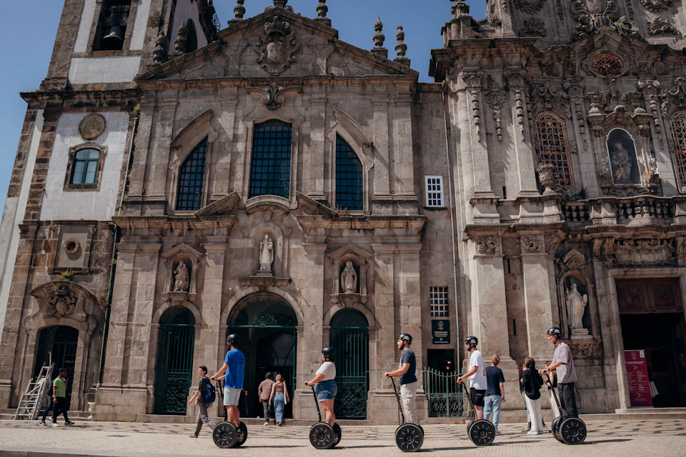 Five people on segways in front of a historic stone building with arched windows and statues. Other pedestrians nearby.