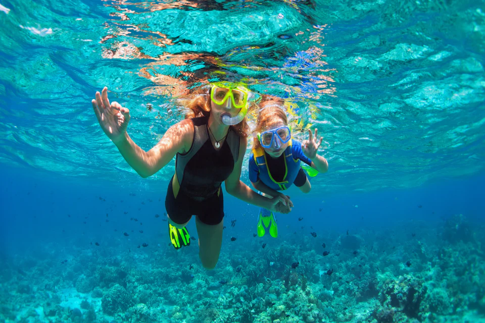 Two people snorkeling in clear blue water above coral reefs, both wearing masks, snorkels, and flippers, making OK signs.