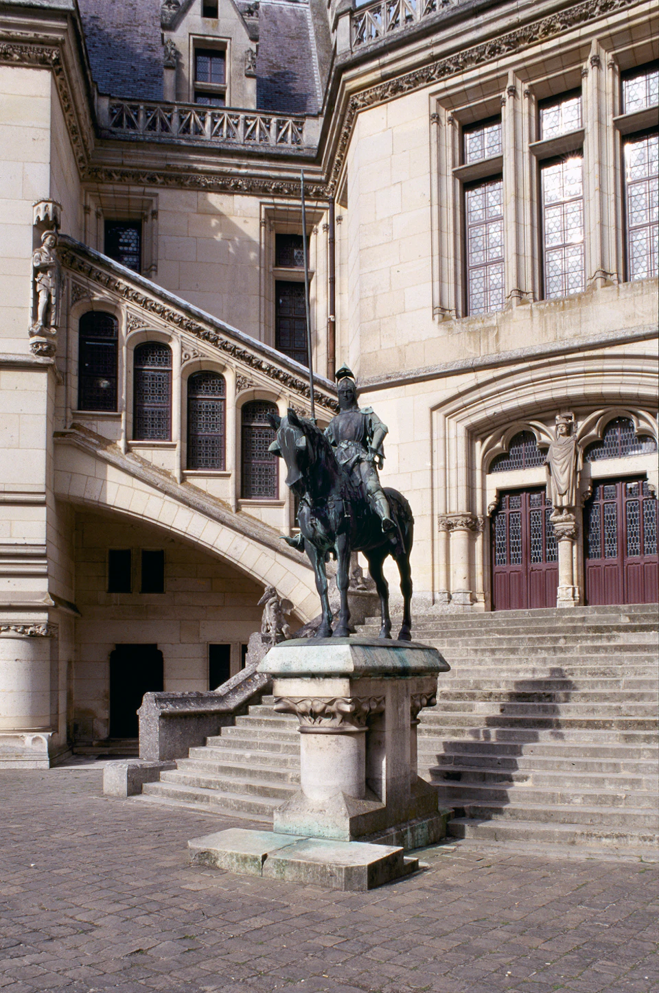 Stone statue of a knight on horseback is positioned in front of an ornate building with stairs, arches, and large windows.