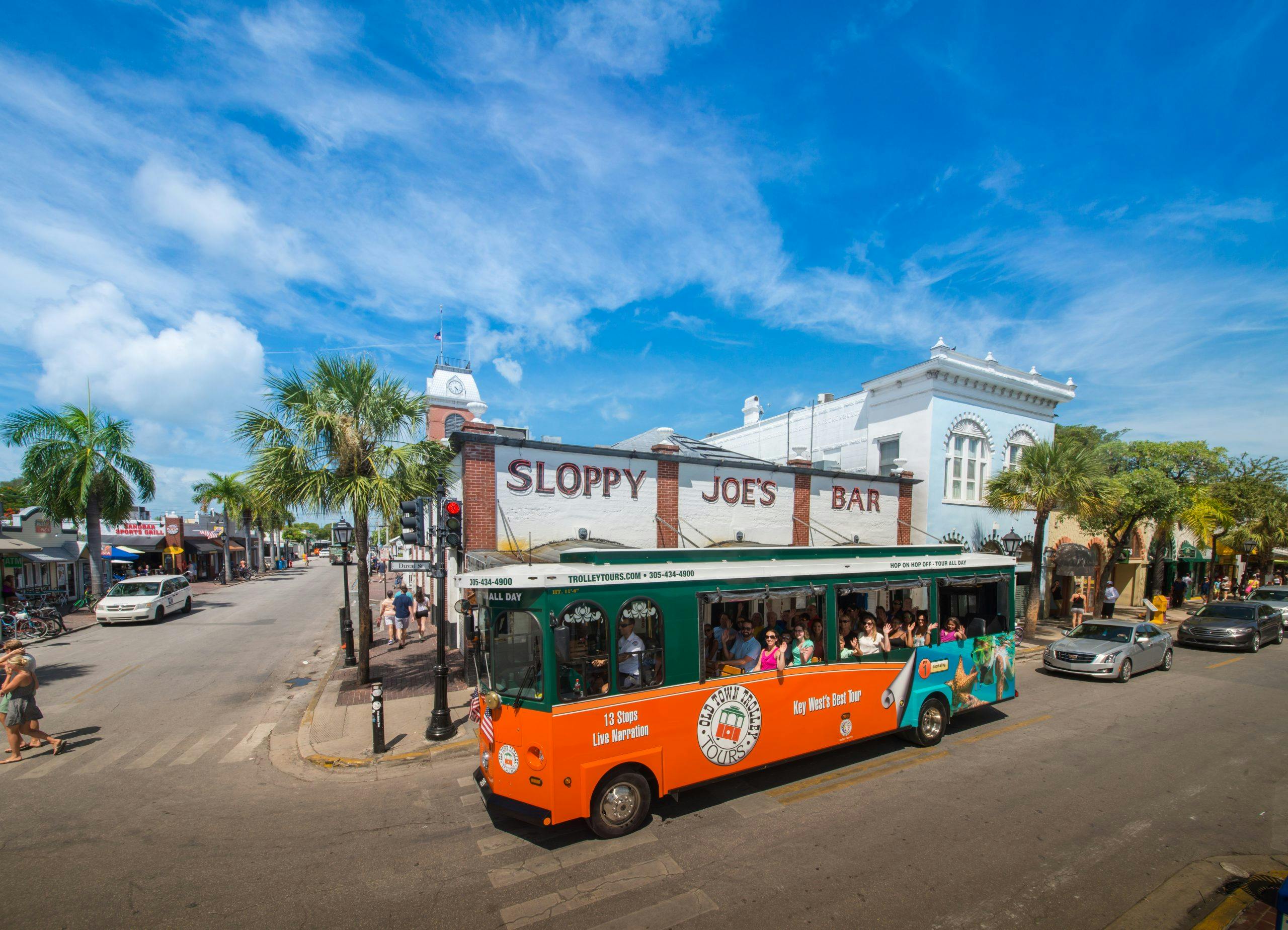 Jasně oranžový a zelený vozík s cestujícími projíždí kolem baru Sloppy Joe's za slunečného dne s několika palmami a modrou oblohou.