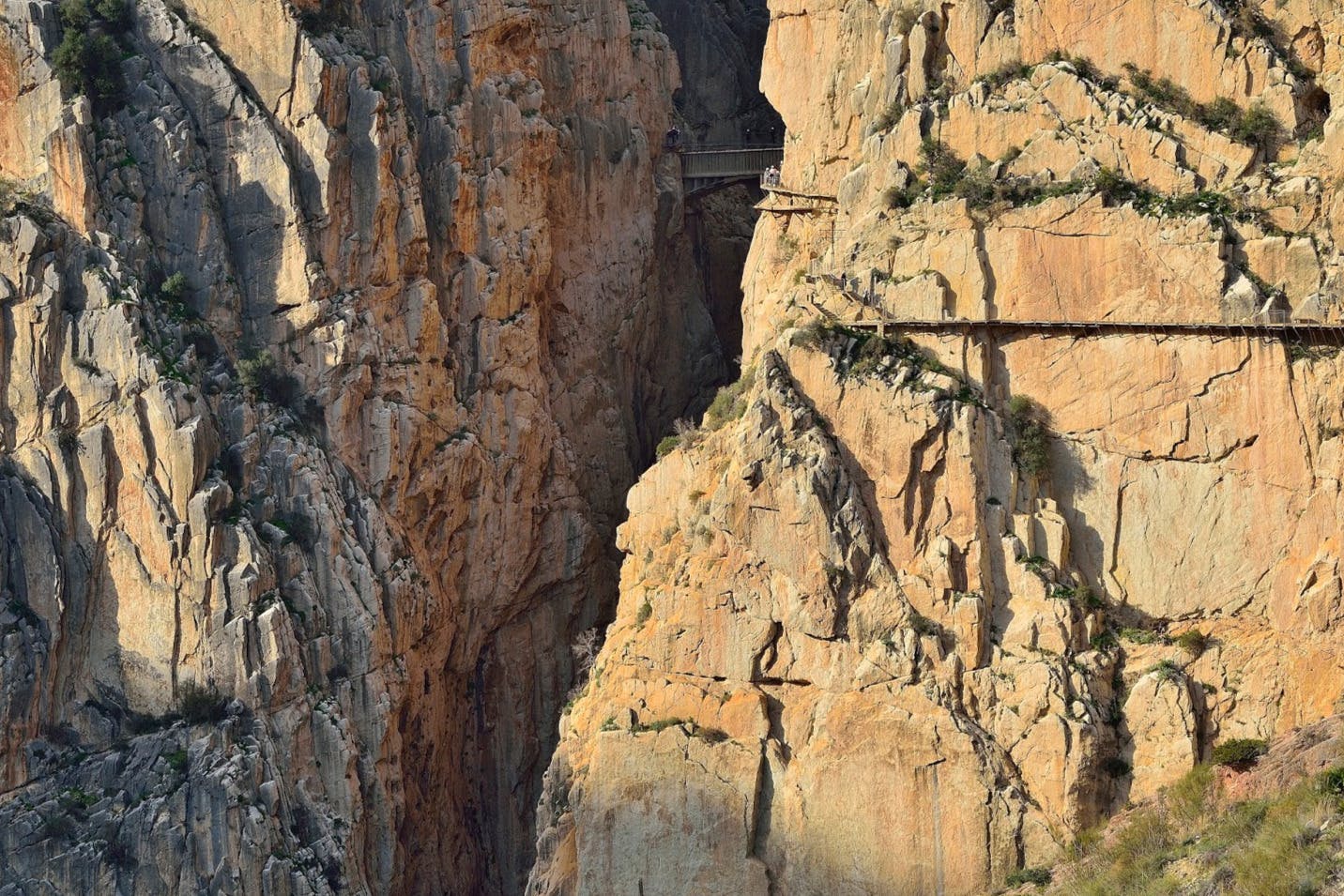 A narrow, rocky canyon with a faint trail and bridge along the steep, rugged cliffs.