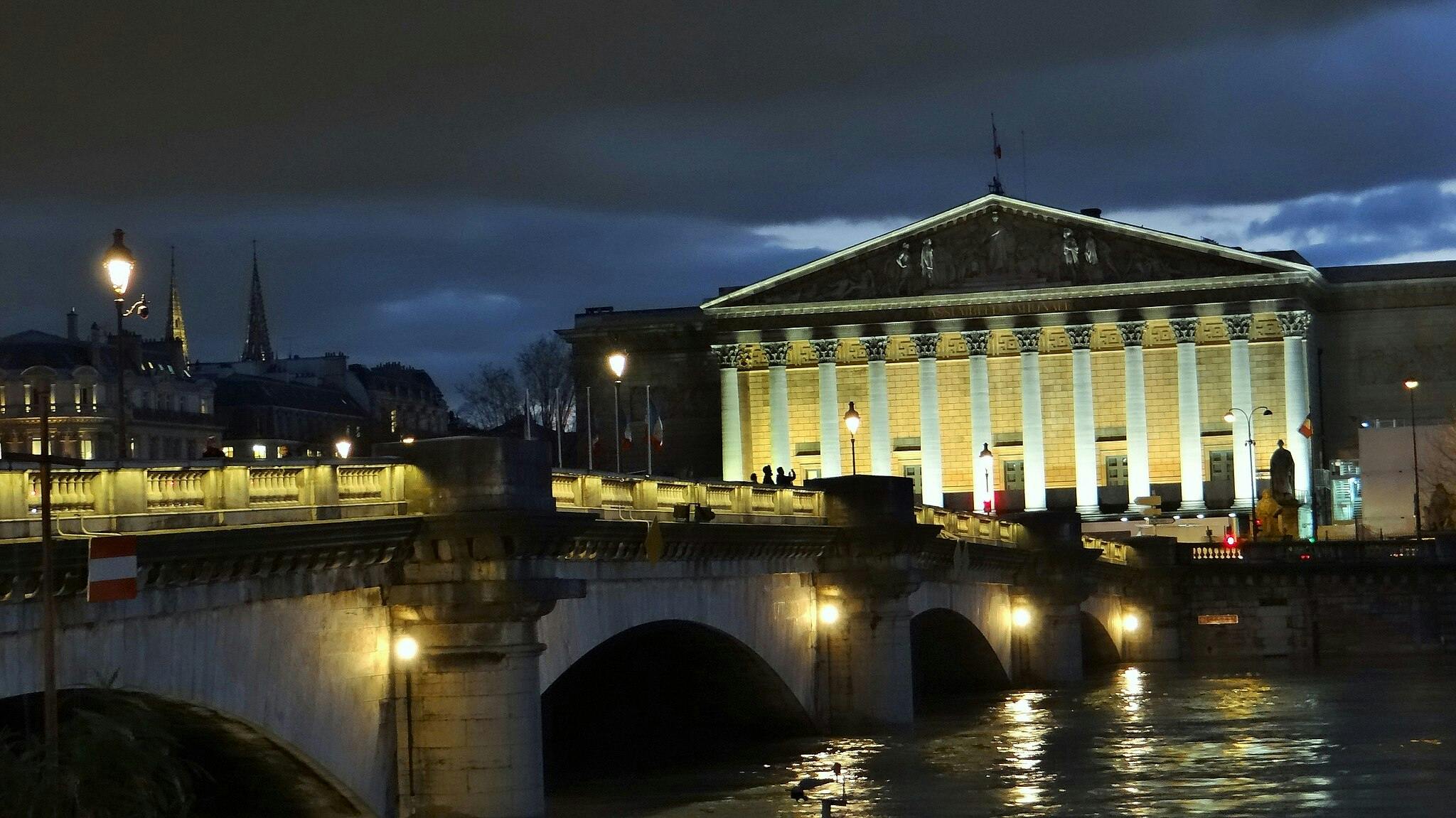 Illuminated bridge and classic building with columns at night, under a cloudy sky and reflections in the water below.