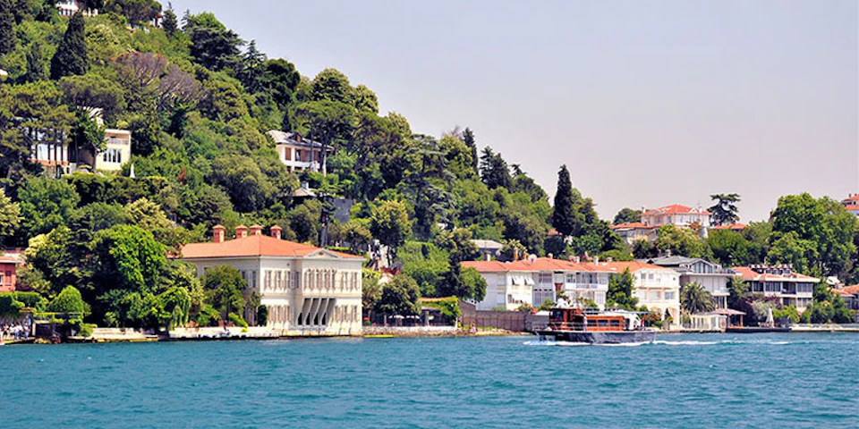 A coastal scene with large waterfront houses and lush greenery on a hillside, and a boat on the blue water in the foreground.