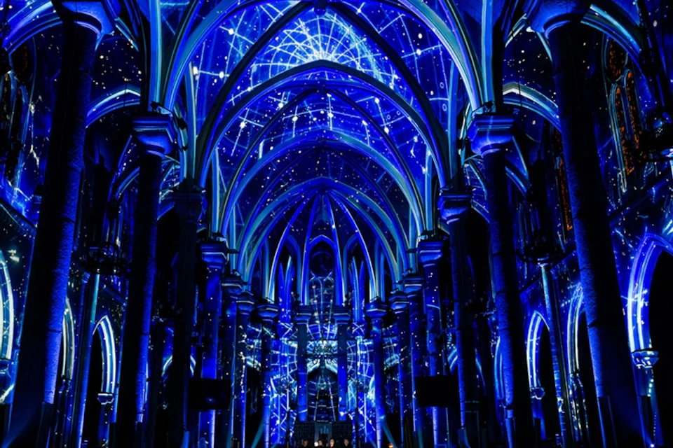 A cathedral interior illuminated with blue light projections resembling constellations and stars on its arches and ceiling.