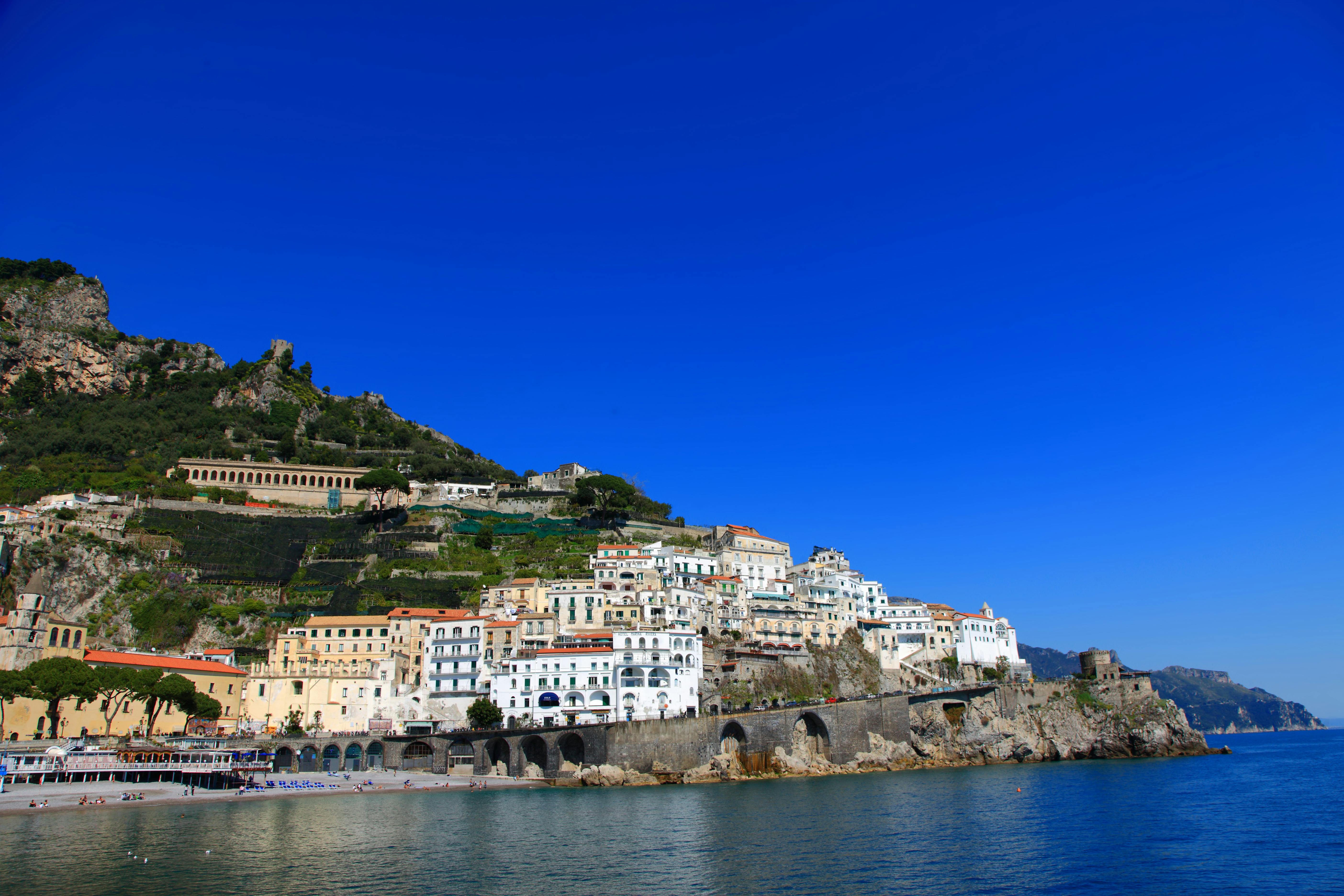 A coastal town with white buildings stacked on a hillside against a clear blue sky, with a calm body of water in the foreground.