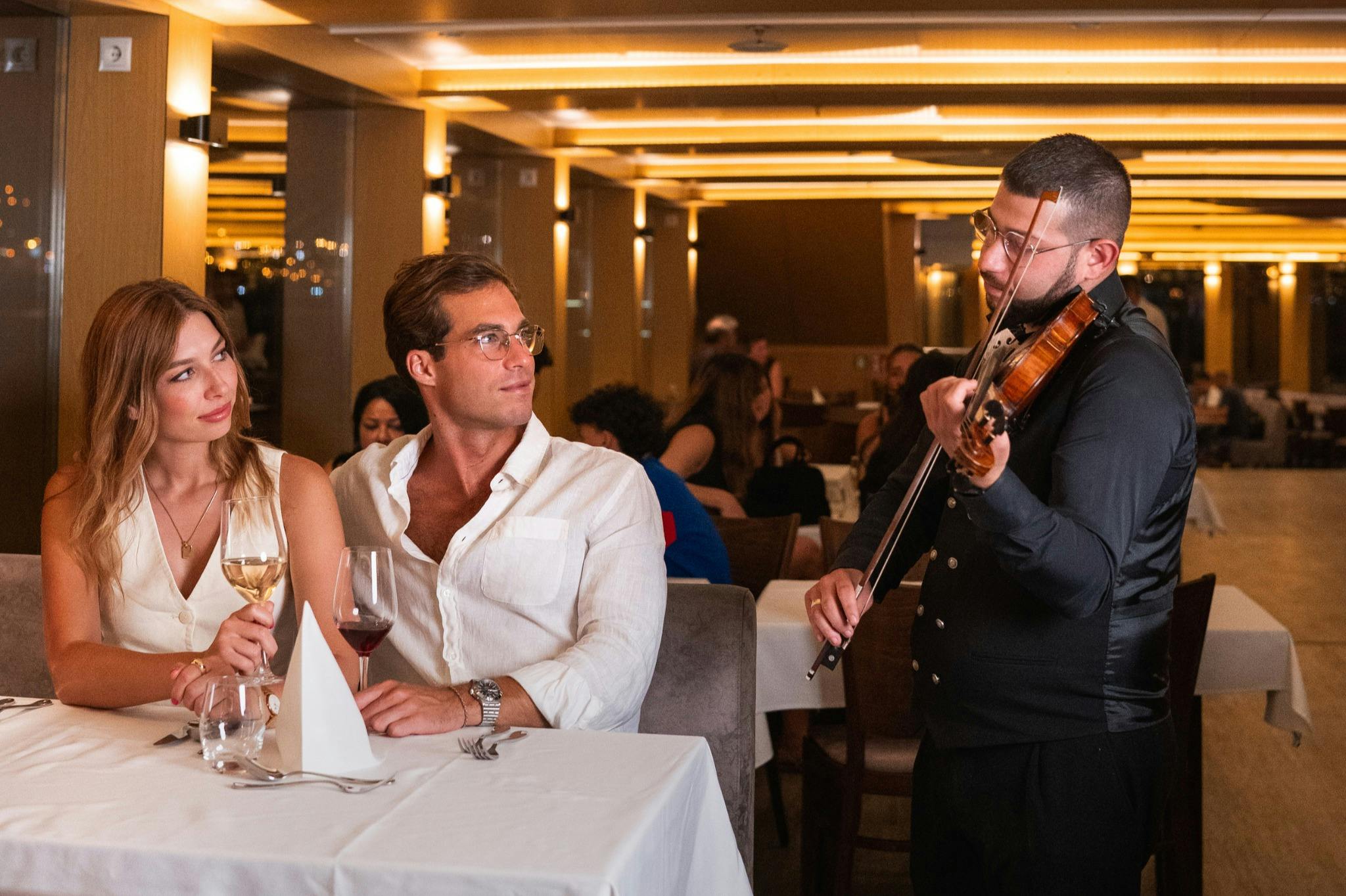 Ein Mann und eine Frau sitzen an einem Restauranttisch, jeder hält ein Glas Wein in der Hand, und im Hintergrund sind andere Gäste zu sehen.