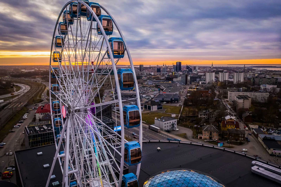 Ferris wheel with blue cabins near a cityscape at sunset. Buildings and streets visible under a cloudy sky.