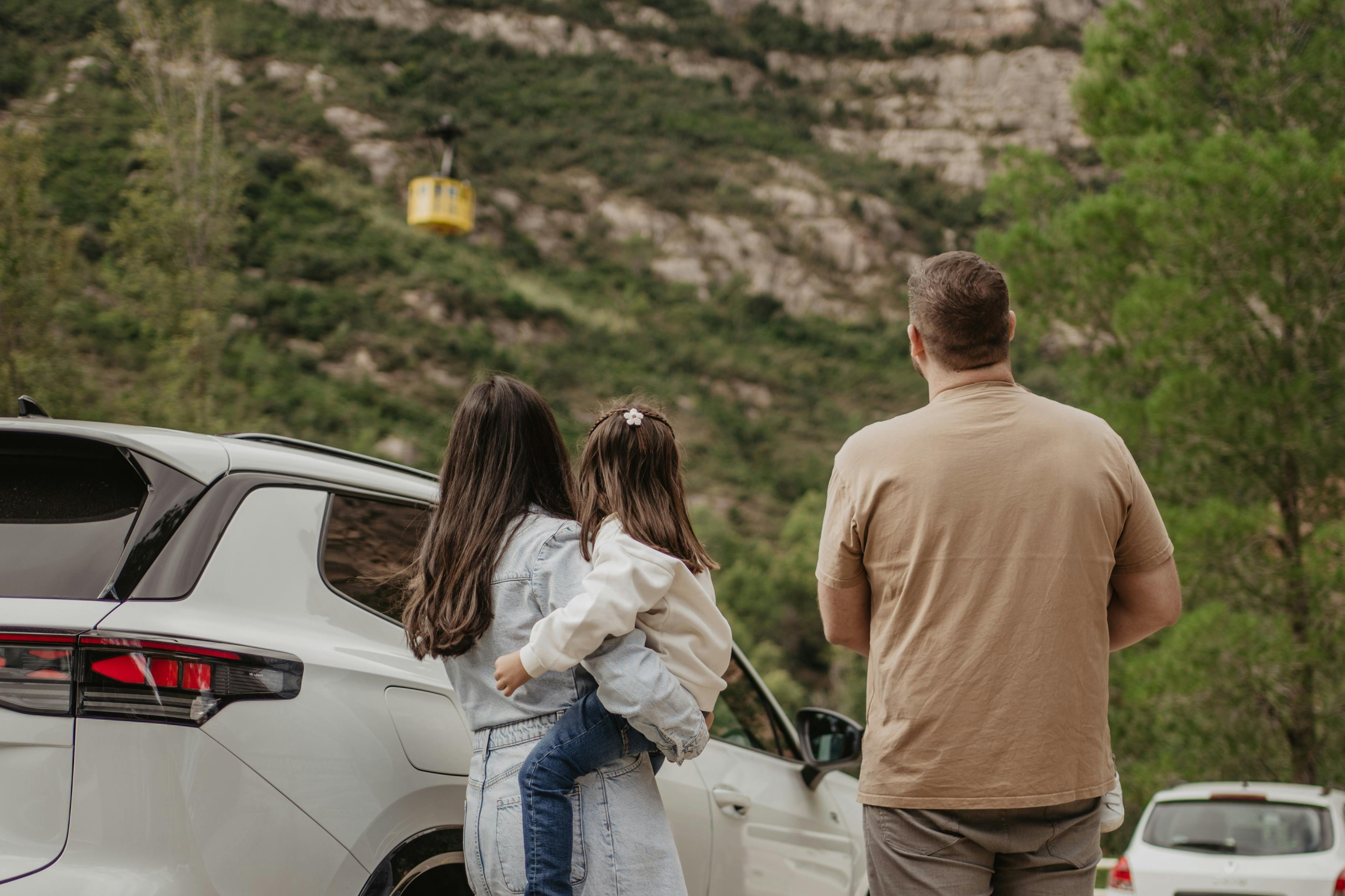 Una familia de tres miembros está de pie cerca de un coche blanco, mirando un teleférico amarillo que asciende por una ladera verde y rocosa.