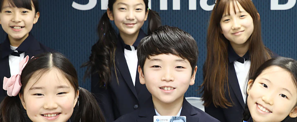 Group of children in uniforms holding cards, smiling in front of a sign with "Shinhan" visible.