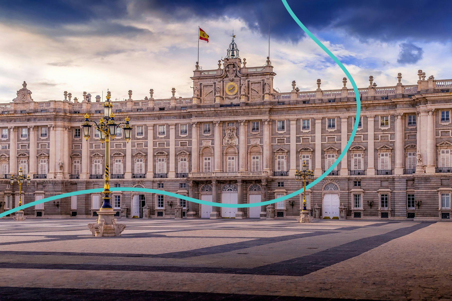 宽敞的宫殿，立面华丽，有多扇窗户，顶部挂着西班牙国旗。 在多云的天空下，前景是空荡荡的庭院。
