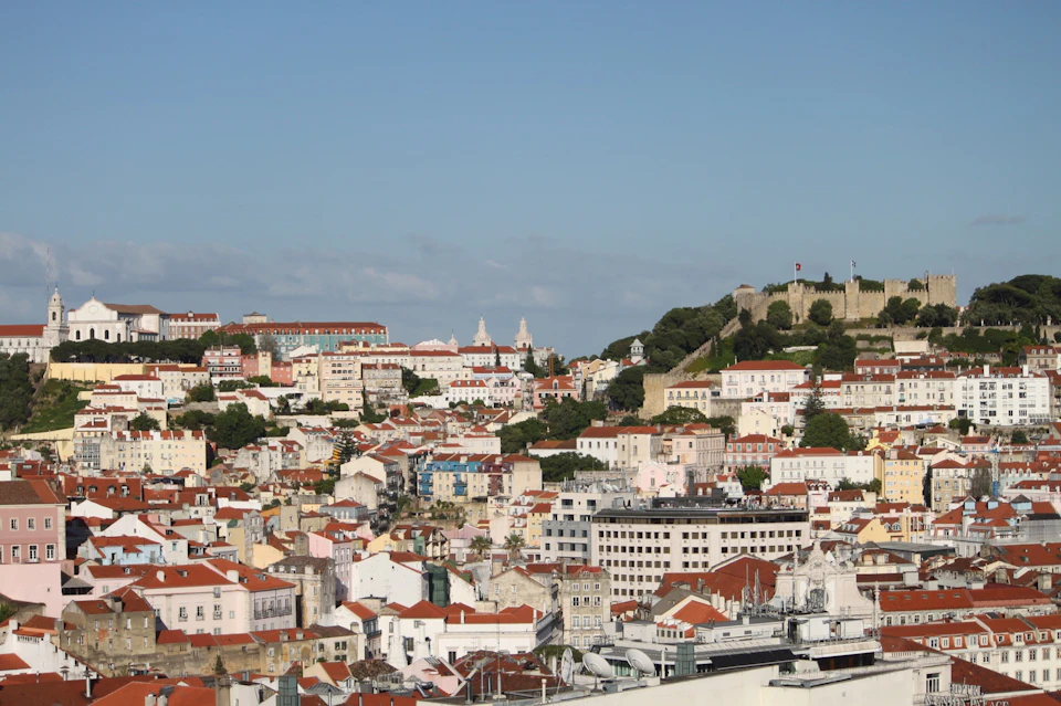 Hillside urban landscape with numerous colorful buildings, a castle with flags, and a clear blue sky.