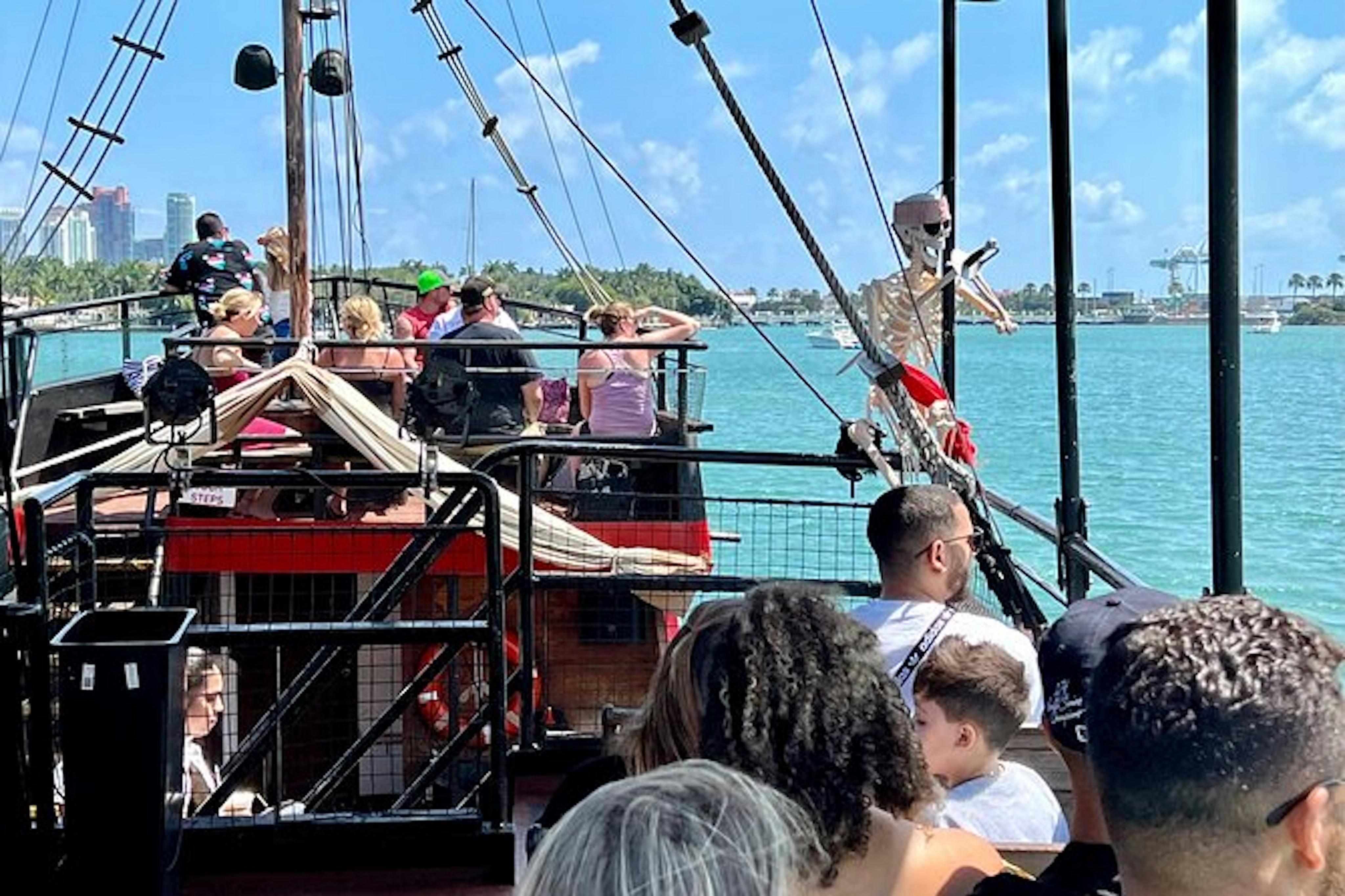 People seated on a boat with a skeleton figure wearing sunglasses attached to its rigging, set against a water and sky background.