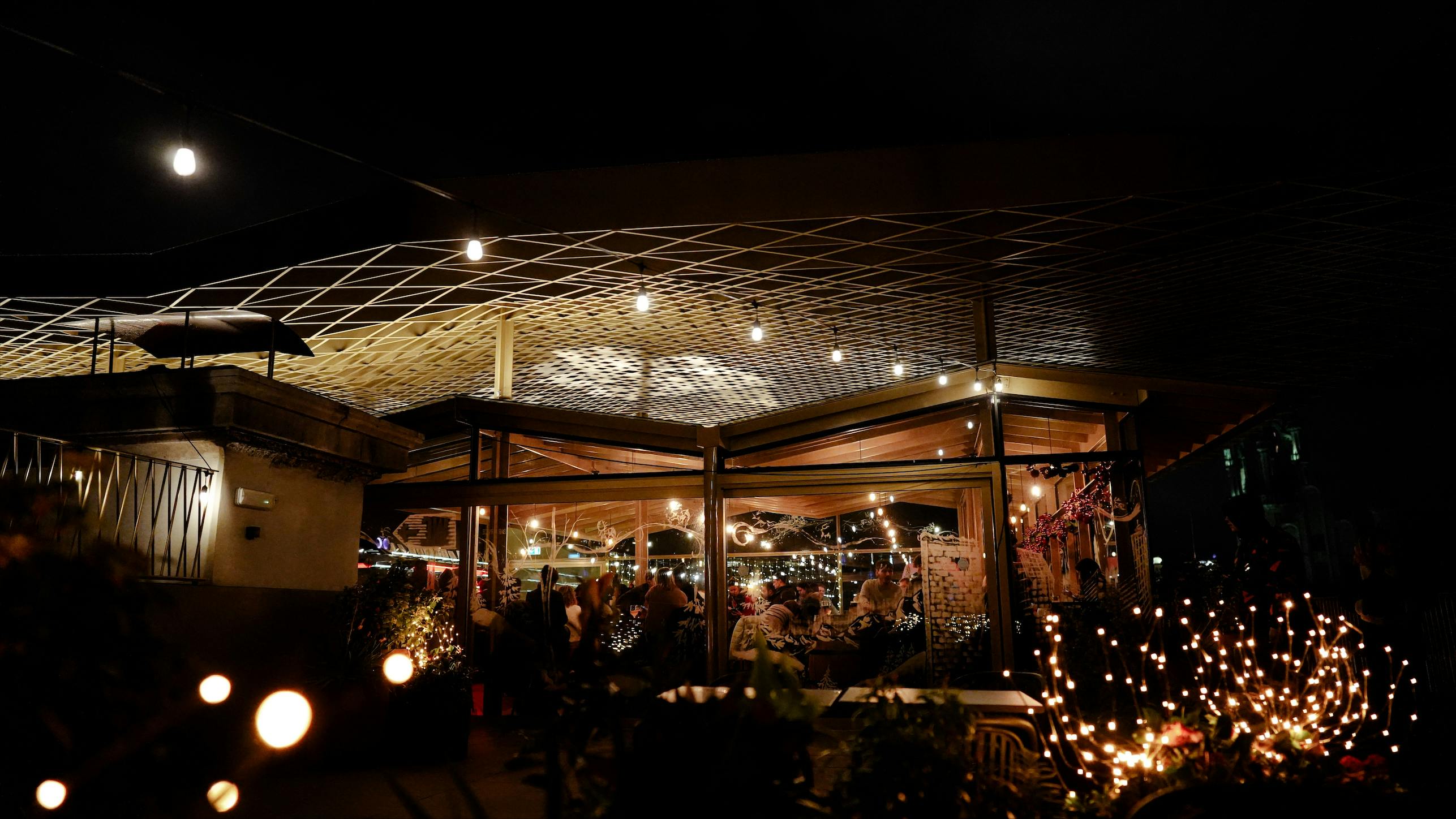 Venkovní restaurace s posezením v noci s provázkovými světly, stoly a lidmi, kteří stolují pod mřížovou střechou.