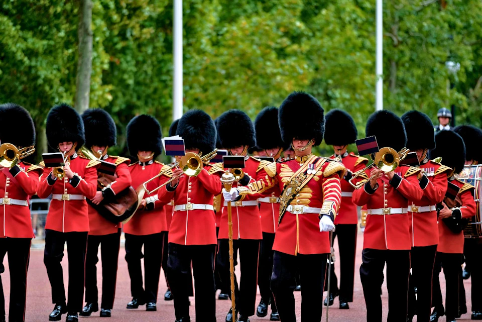 A marching band in red uniforms and black bearskin hats performing outdoors with musical instruments.