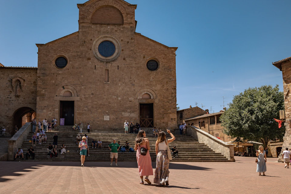 People walking and sitting near a historic brick church with circular windows, under a clear blue sky.