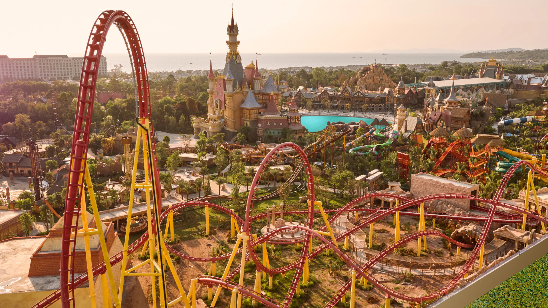 A theme park with a red and yellow roller coaster, a castle-like structure, and other attractions under a sunny sky.