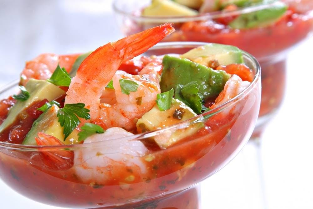 Glass bowl of shrimp cocktail with avocado, tomatoes, and parsley, served in a bright and fresh presentation.