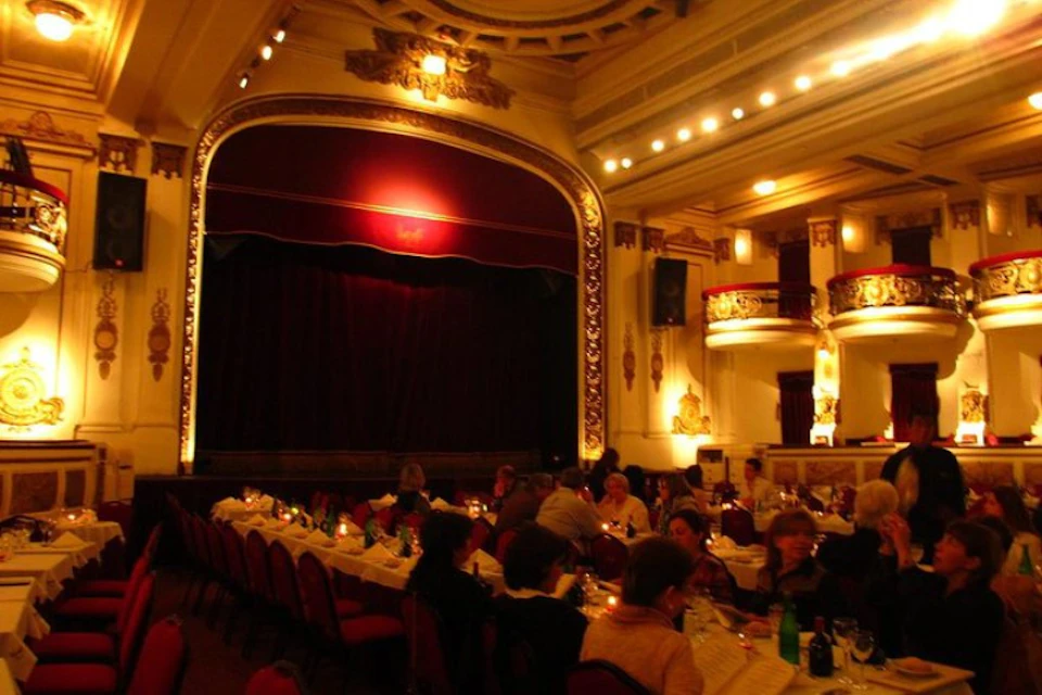Dimly lit theater with red chairs, ornate walls, and a stage with red curtains. People are dining at tables with candlelight.