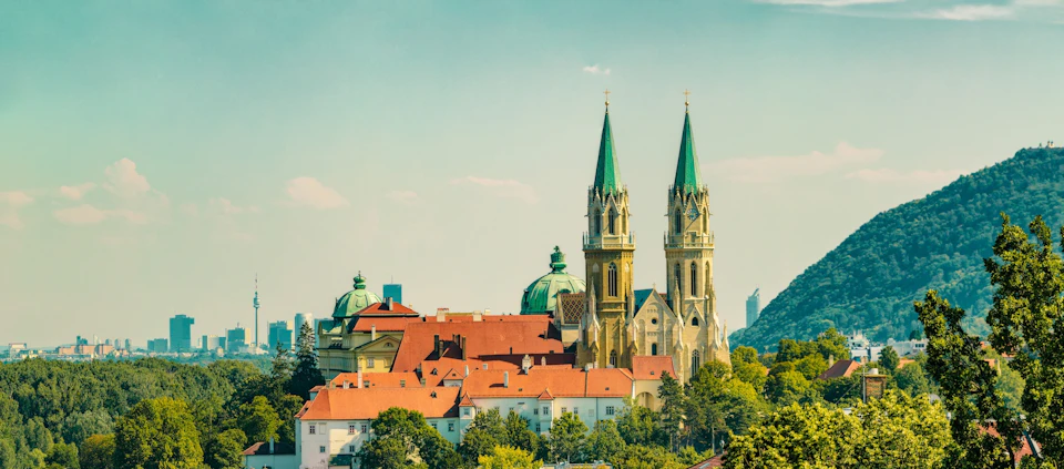 A scenic view of a town with a church featuring twin spires, red-roofed buildings, and a backdrop of trees and blue sky.