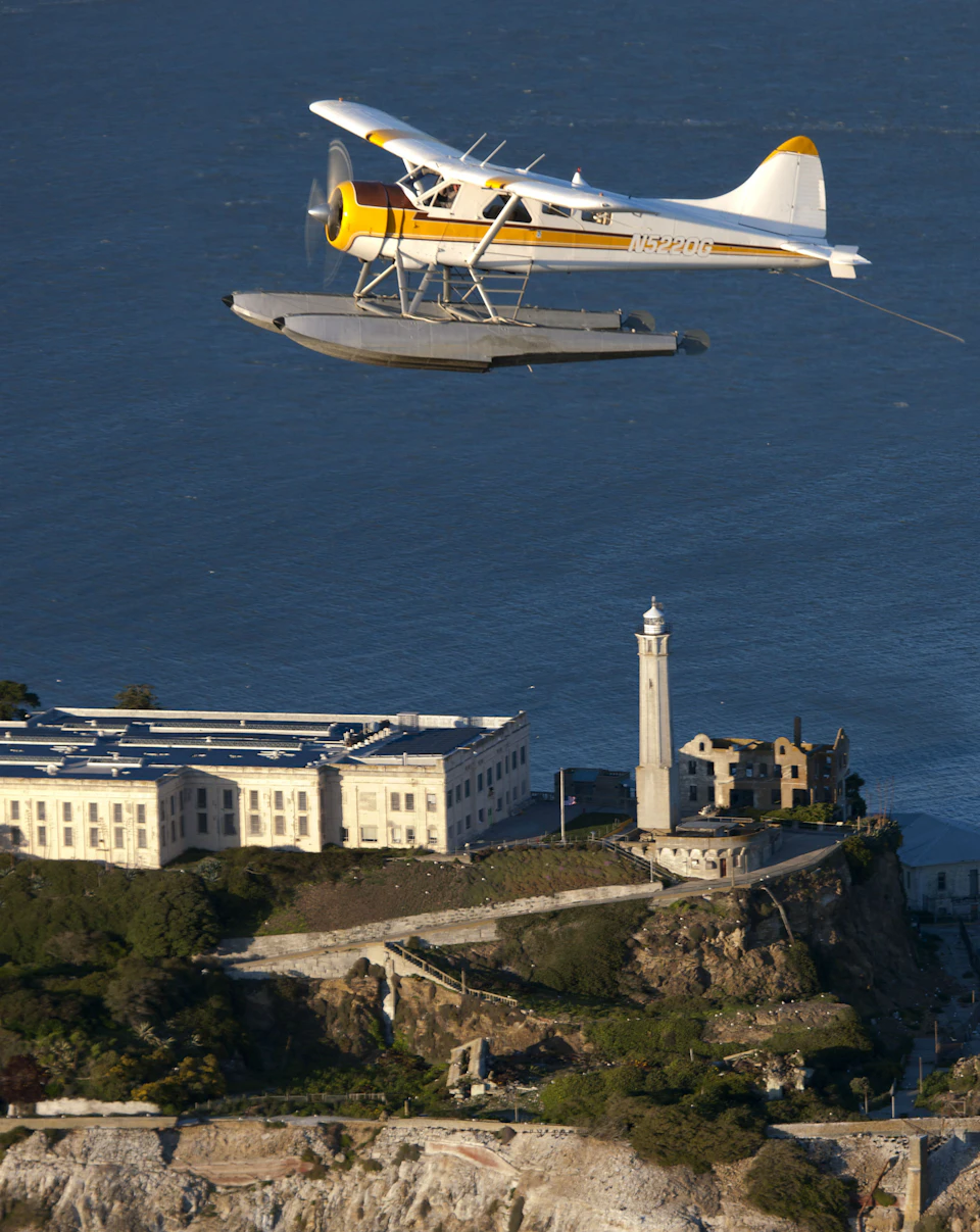A seaplane flies over a coastal fortress with a tall lighthouse and a large building by the water.
