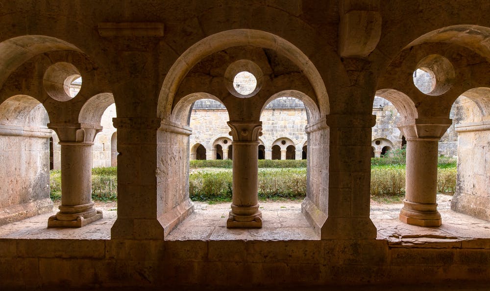 Μια πέτρινη καμάρα με τρία ανοίγματα πλαισιώνει μια αυλή με πράσινο και ένα παλιό κτίριο με τοξωτά παράθυρα στο παρασκήνιο.