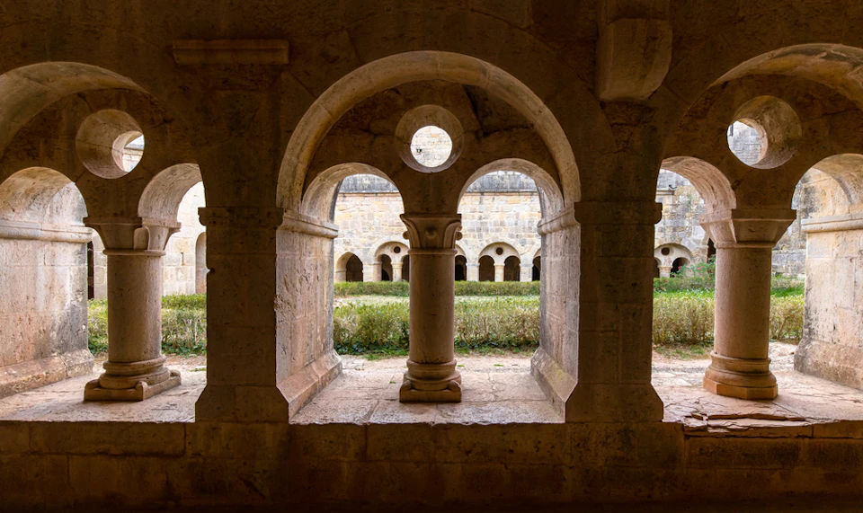 A stone archway with three openings frames a courtyard with greenery and an old building with arched windows in the background.
