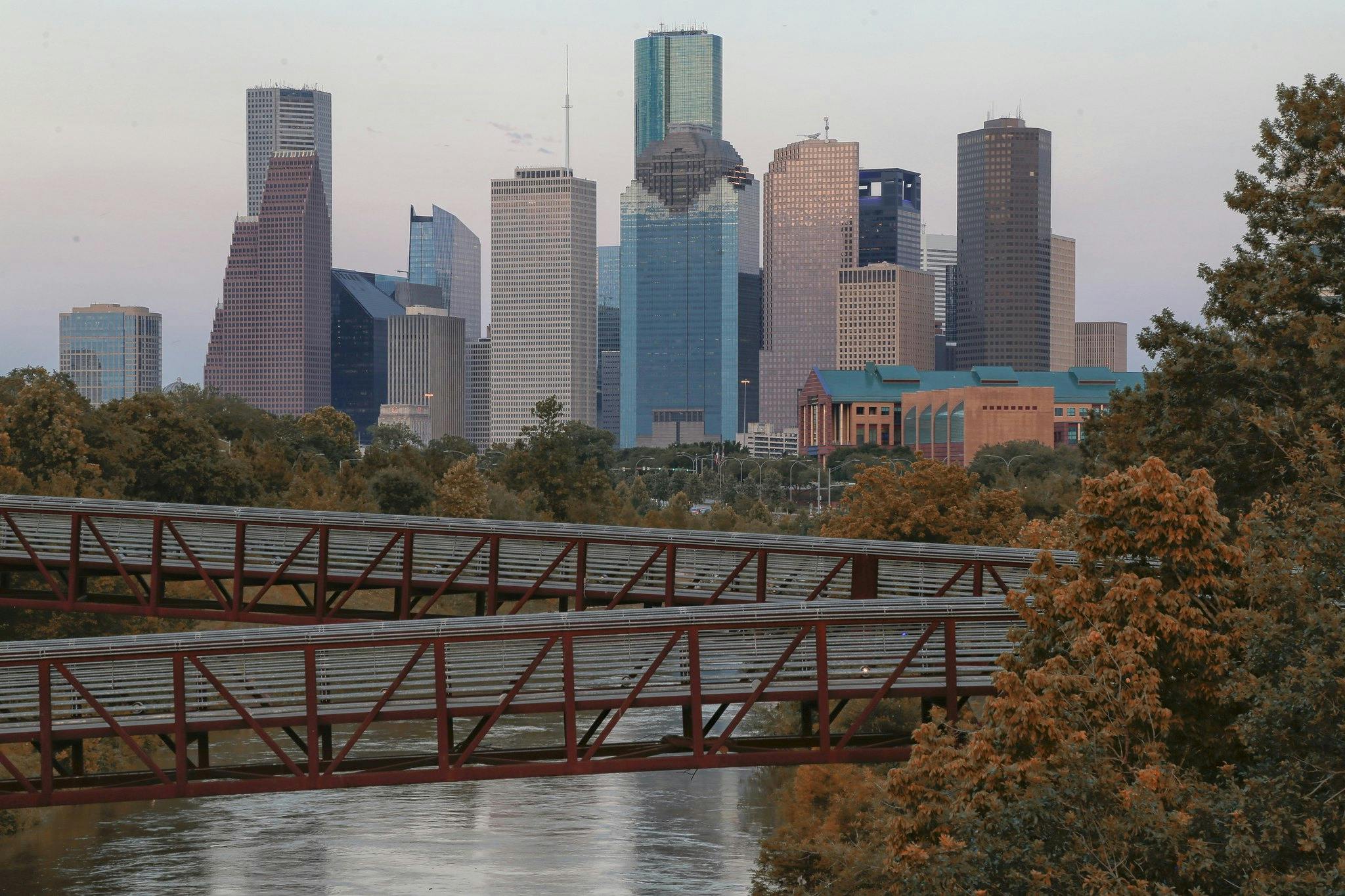 Panorama miasta z wysokimi budynkami i mieszanką projektów architektonicznych. Drzewa i dwa metalowe mosty nad rzeką wypełniają pierwszy plan.