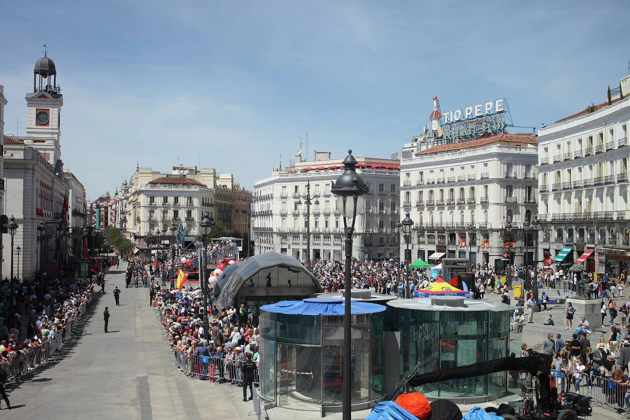 Uma grande multidão reúne-se numa praça da cidade rodeada de edifícios históricos, incluindo uma torre de relógio e o letreiro Tio Pepe.