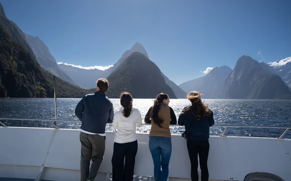 Four people stand on a boat's railing, overlooking a scenic fjord with mountains under a clear blue sky.