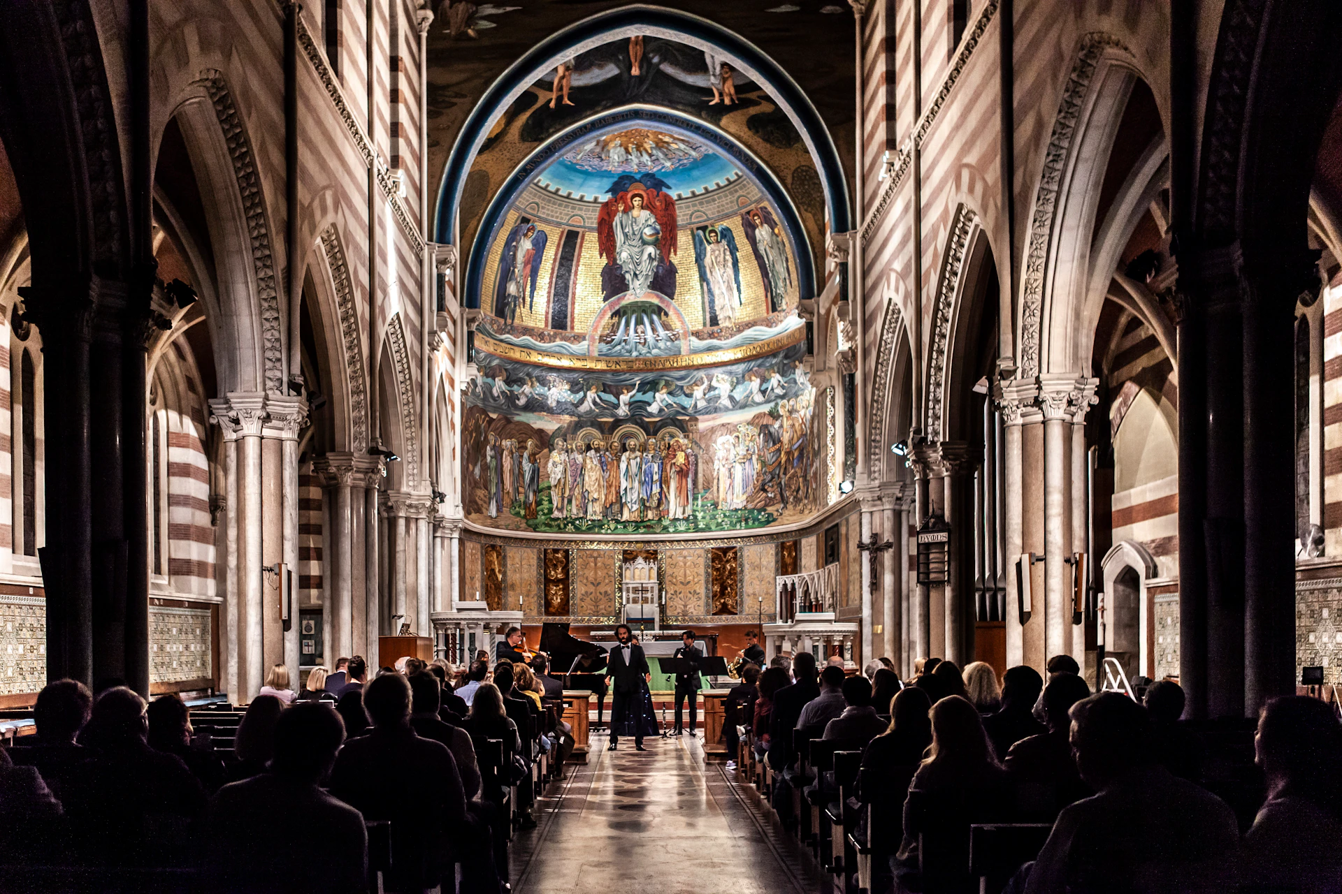 A choir performs in a grand cathedral with an elaborately painted dome, flanked by columns and an audience sitting in pews.