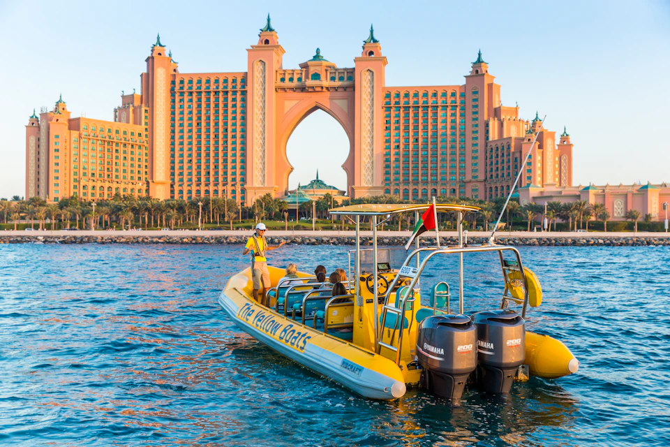 A yellow boat with people onboard is near water, in front of a grand, arch-shaped building with towers and palm trees.