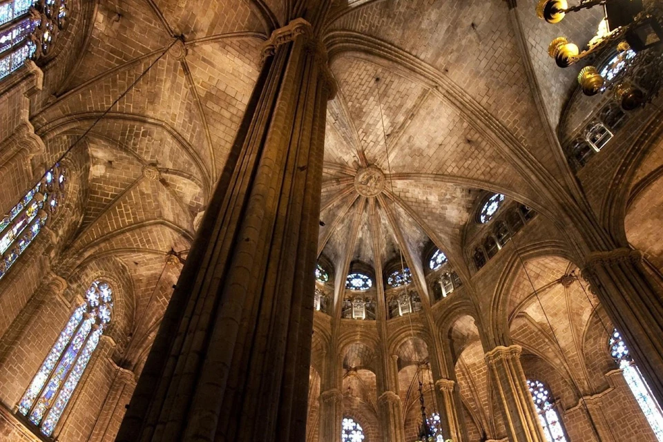 Interior view of a gothic cathedral's vaulted ceiling, showing intricate stonework, columns, and stained glass windows.