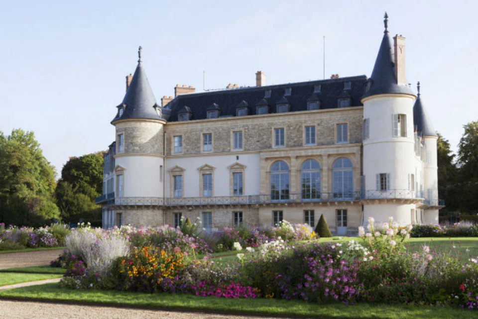 A grand chateau with multiple arched windows, towers, and a manicured garden filled with colorful flowers in the foreground.
