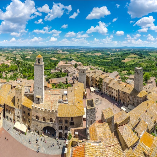 Panoramic view of a historic town with stone buildings and towers, surrounded by a lush green landscape under a bright blue sky with clouds.