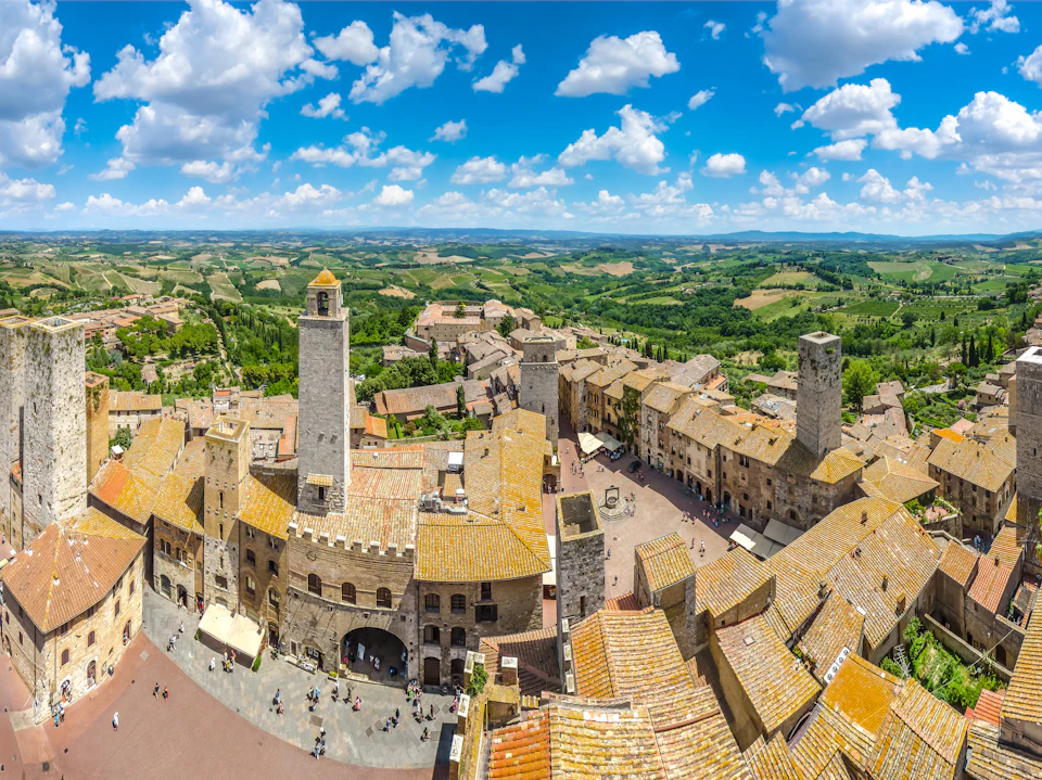 Panoramic view of a historic town with stone buildings and towers, surrounded by a lush green landscape under a bright blue sky with clouds.